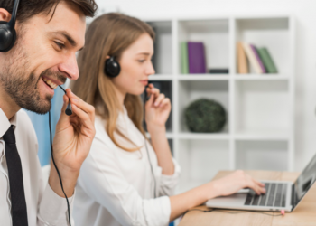 Imagem de dois vendedores, um homem e uma mulher com head sets falando e olhando para a tela de seus computadores.
