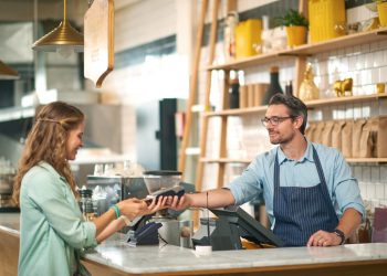 Vendedor no caixa de uma mercearia segura uma máquina de cartão para que a cliente possa digitar sua senha