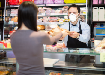 Vendedor atrás do balcão usando máscara entrega uma mercadoria para cliente.