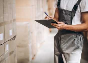Homem olha para caixas em um estoque e anota algo em sua prancheta