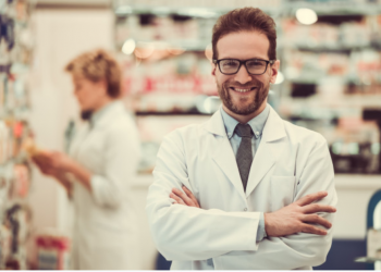 Homem de jaleco e braços cruzados olhando para a câmera e sorrindo dentro de uma farmácia